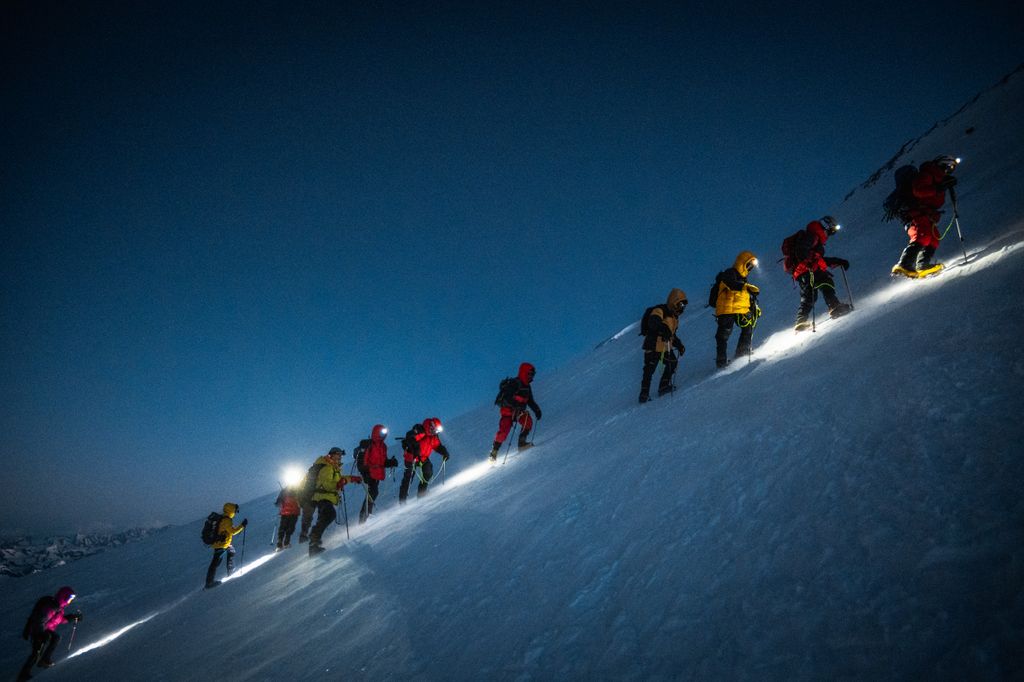 A group of climbers ascend Mount Elbrus at night, guided by headlamps, on a steep snowy slope.