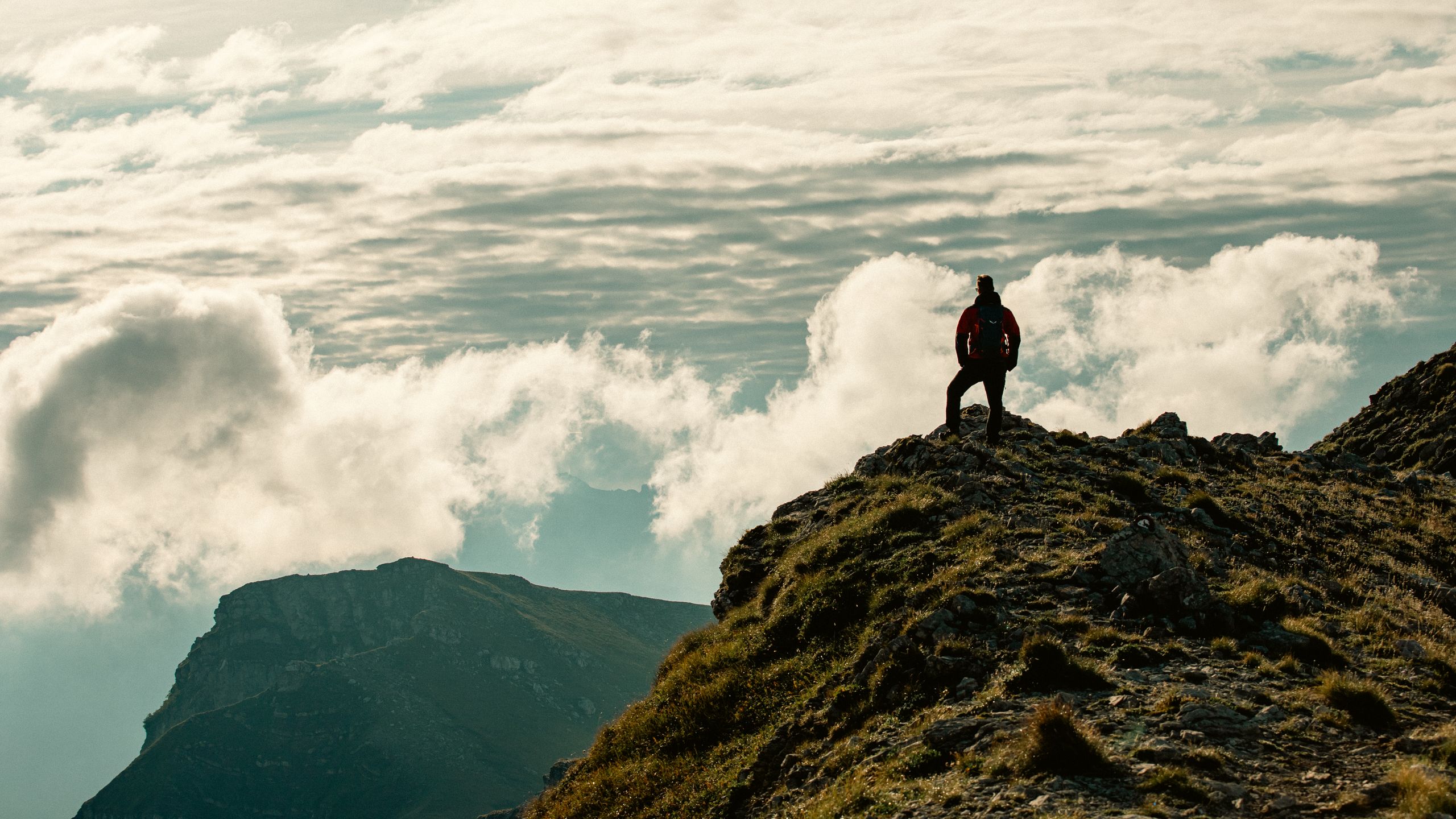 A lone hiker stands on a mountain peak, surrounded by breathtaking clouds and rugged terrain.