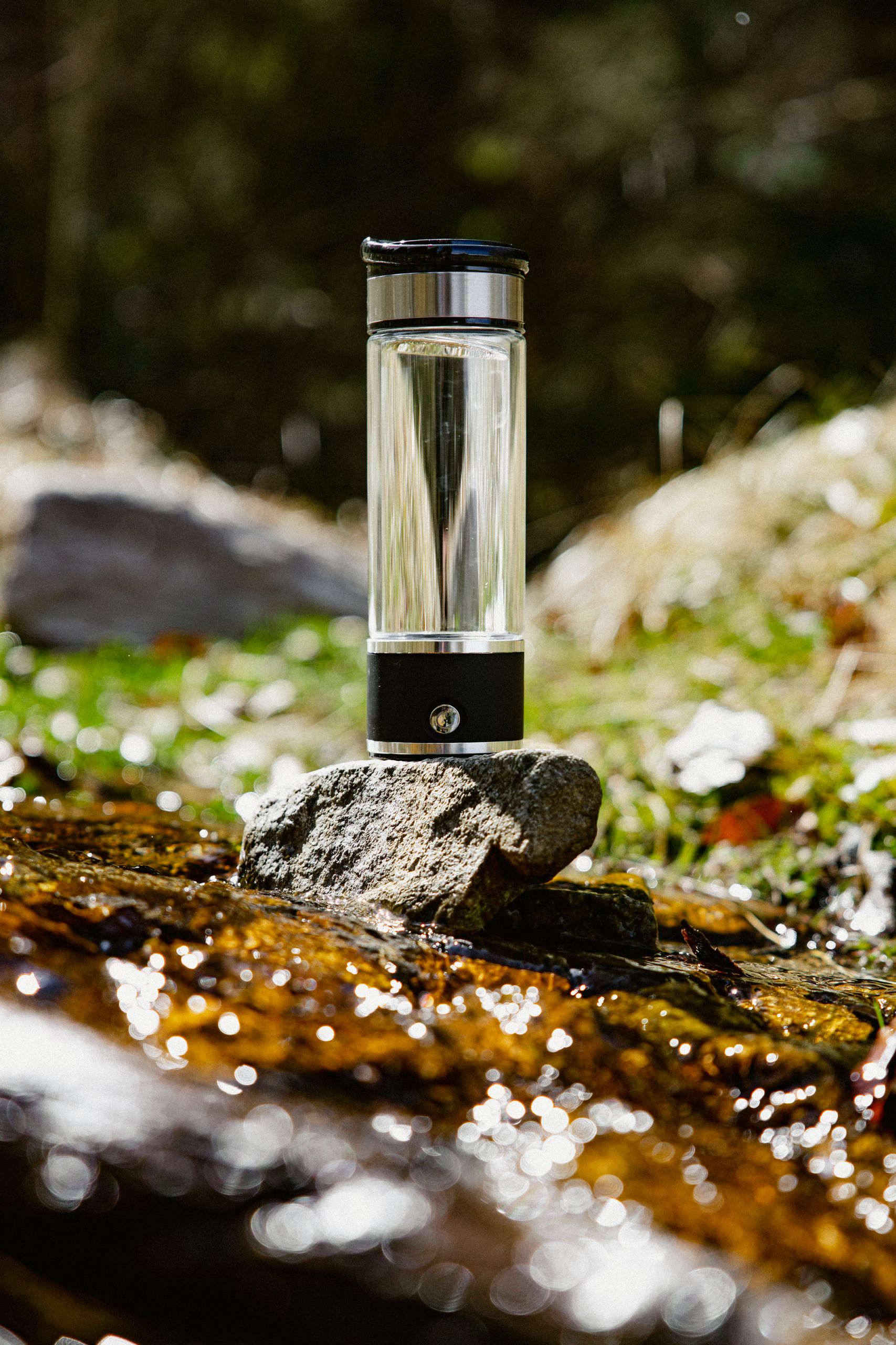 A sleek glass water bottle sits on a rock by a stream, highlighting purity and nature.