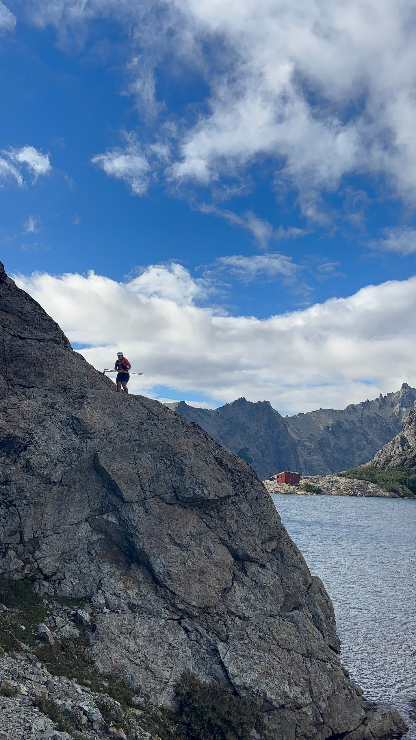 A solitary hiker stands atop a rocky cliff overlooking a serene lake under a clear blue sky.