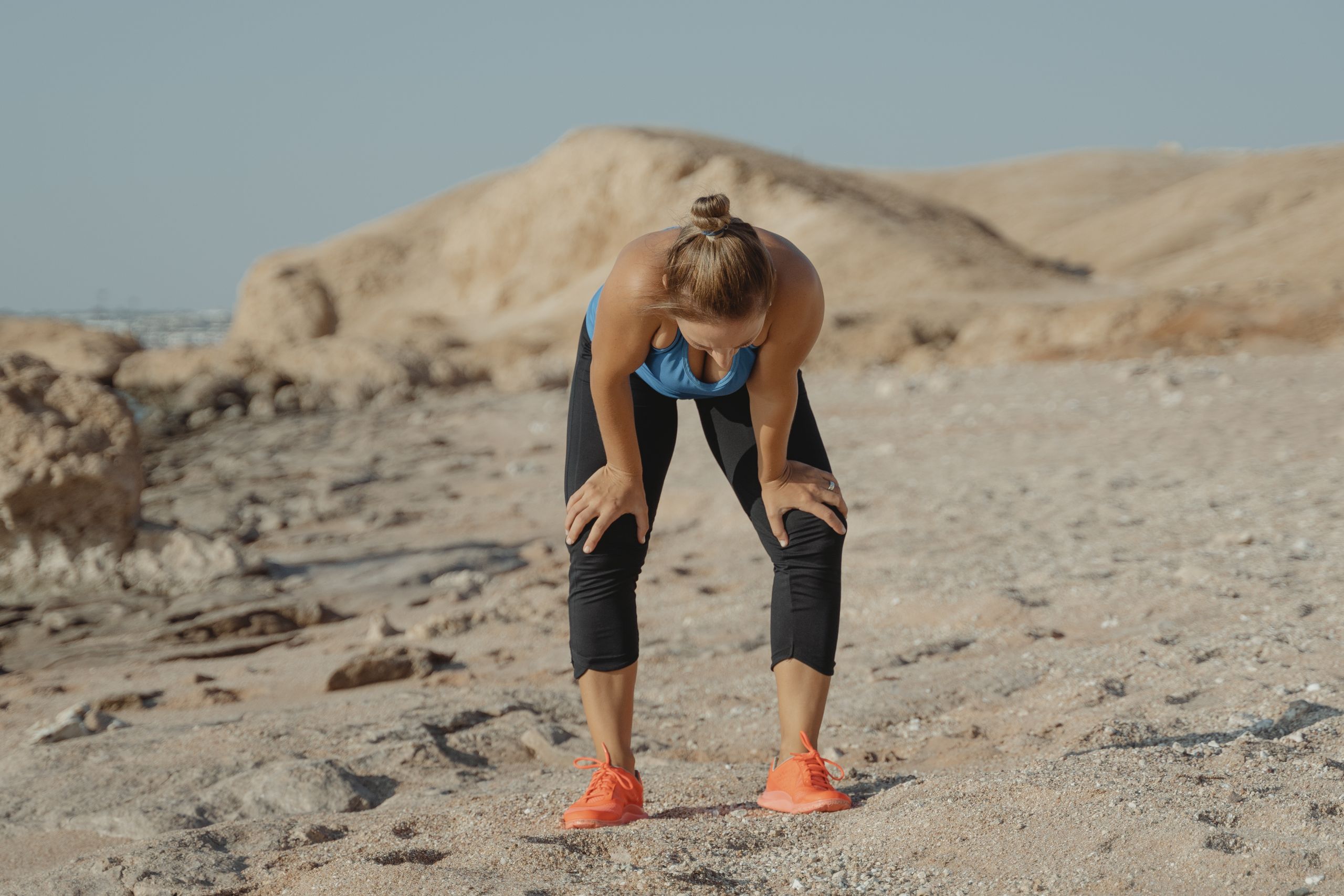 A tired woman in athletic wear taking a break on a sandy beach under a clear sky.