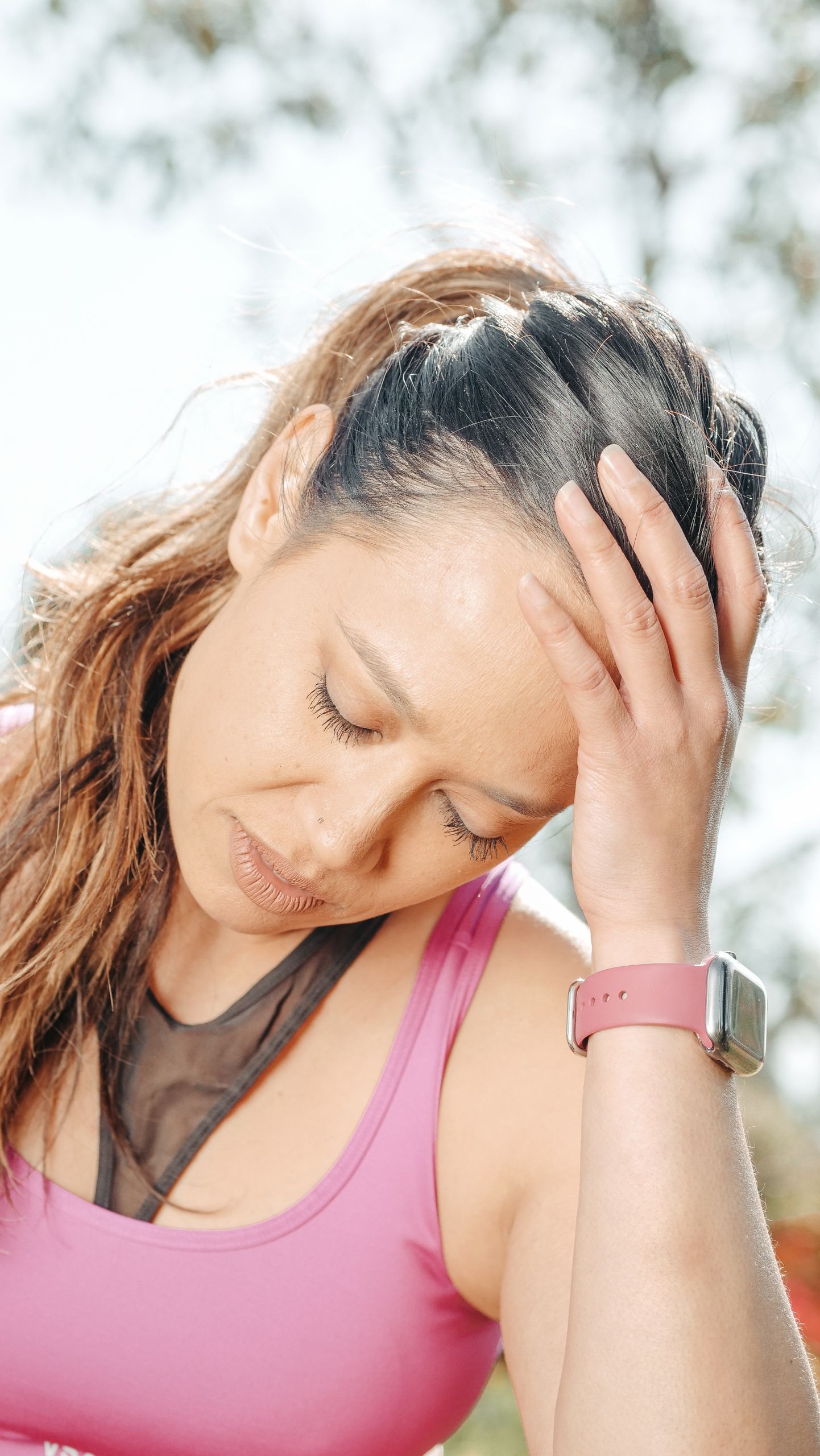 A woman in a pink tank top experiencing a headache while outdoors, holding her head in pain.