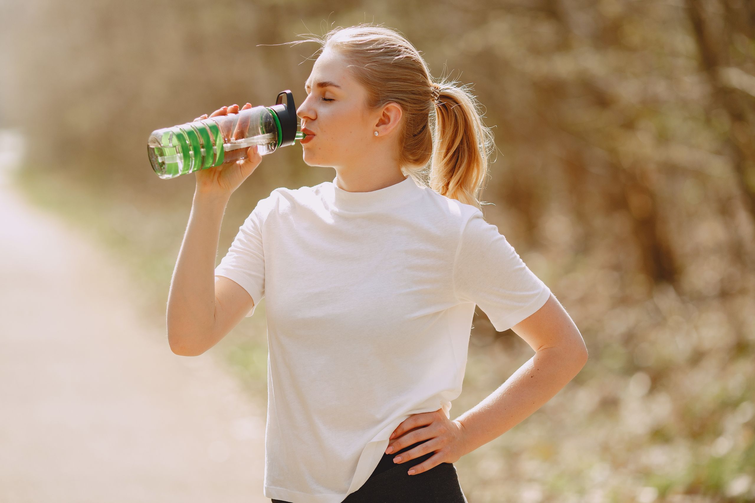 A young woman in a white shirt staying hydrated outdoors with a water bottle in sunlight.