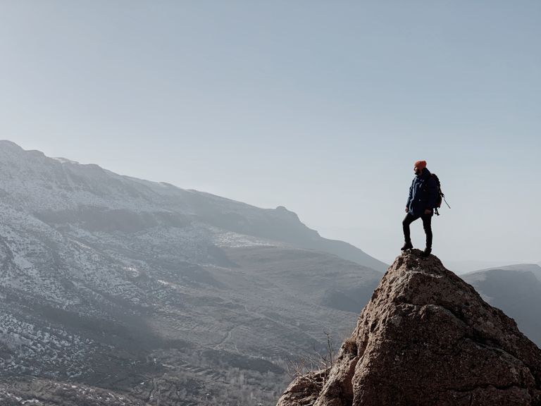 An adventurous solo hiker stands on a rocky peak, overlooking vast mountainous terrain at sunrise.