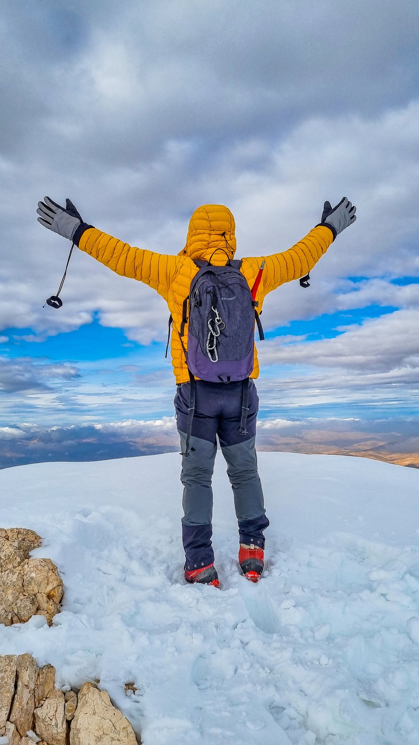 Man in yellow jacket celebrating atop snowy mountain peak during winter hike.