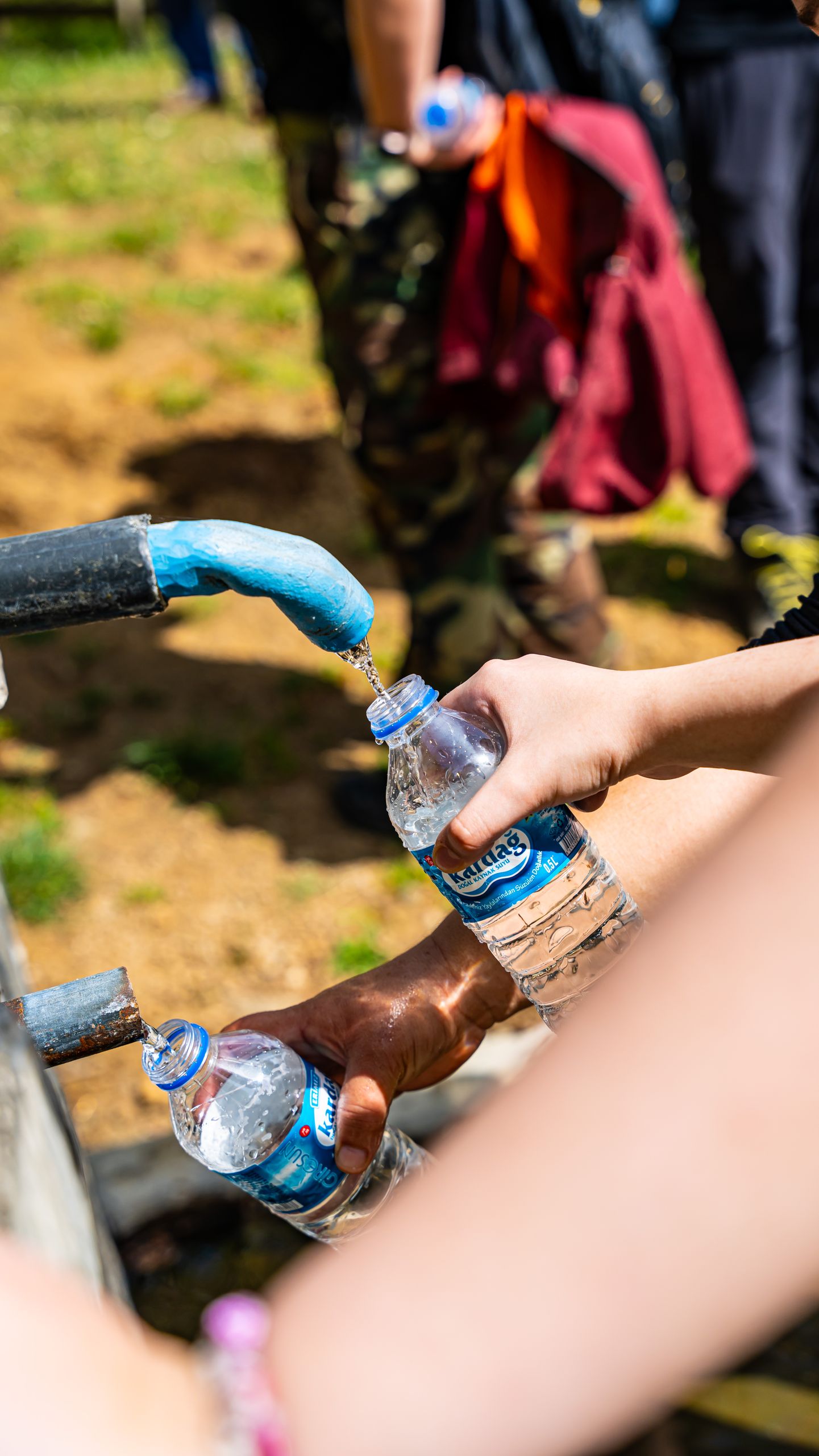 People filling water bottles from an outdoor tap in Düzköy, Trabzon, Türkiye, on a sunny day.